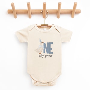 May include: A cream-colored baby onesie with a whimsical goose graphic wearing a party hat and bow. The word "ONE" is in blue block letters, with "silly goose" below. The onesie hangs on a wooden hanger.