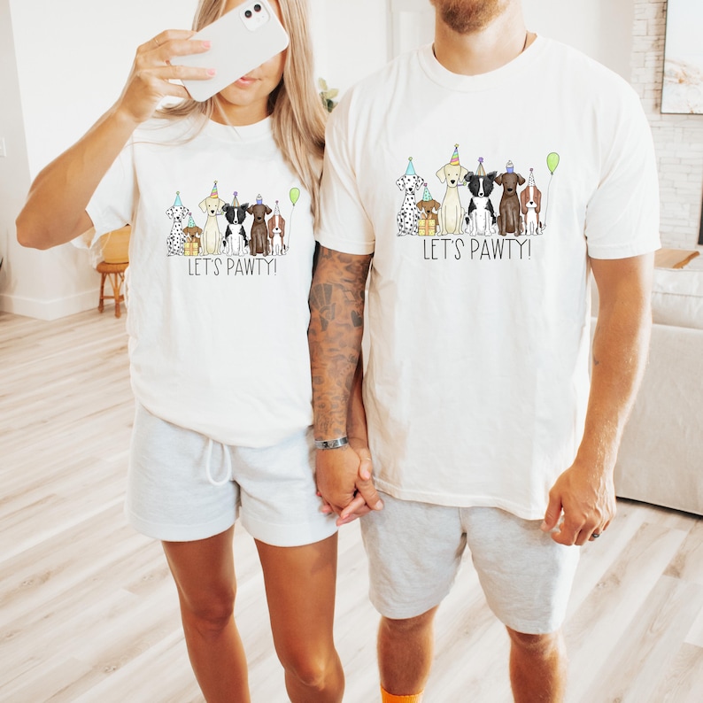 Op de afbeelding: Een stel draagt witte T-shirts met een afbeelding van honden die feestmutsen dragen en de tekst "Let's Pawty!" Het stel houdt elkaars hand vast en glimlacht.
