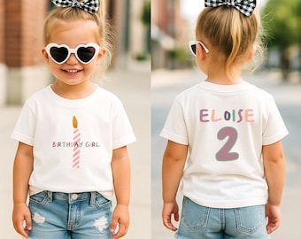 May include: A white t-shirt with a birthday candle graphic and the words "BIRTHDAY GIRL" in pastel colors. The back of the shirt has the name "ELOISE" and the number "2". The child is wearing denim shorts and heart-shaped sunglasses.