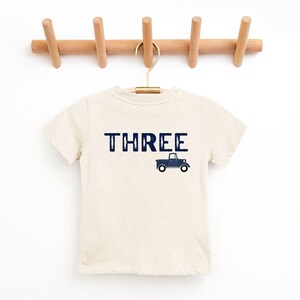 May include: A beige short-sleeved t-shirt with the word "THREE" in navy blue block letters and a navy blue truck graphic. The shirt is hanging on a wooden rack with a gold hanger.