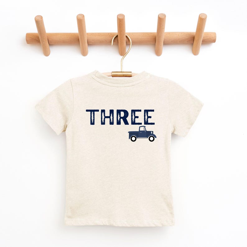 May include: A beige short-sleeved t-shirt with the word "THREE" in navy blue block letters and a navy blue truck graphic. The shirt is hanging on a wooden rack with a gold hanger.