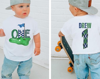 May include: A white t-shirt with a green golf course design and the word "ONE" in blue and white plaid. The number "1" is in the center of the word "ONE" and is decorated with white polka dots. A blue flag is on a golf club in the upper right corner of the design. The back of the shirt has the name "DREW" in blue and white plaid and the number "1" in green and blue plaid.