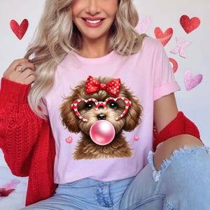 Puede incluir: Una camiseta rosa con un perro marrón que lleva gafas en forma de corazón y un lazo rojo con lunares blancos. El perro está soplando una burbuja rosa.