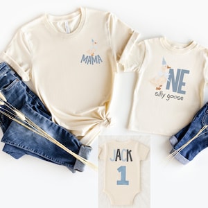 May include: Cream-colored t-shirts and a onesie with blue text and graphics. One shirt says "MAMA" and features a goose graphic. Another shirt says "ONE silly goose". The onesie says "JACK 1". Blue jeans are also in the image.
