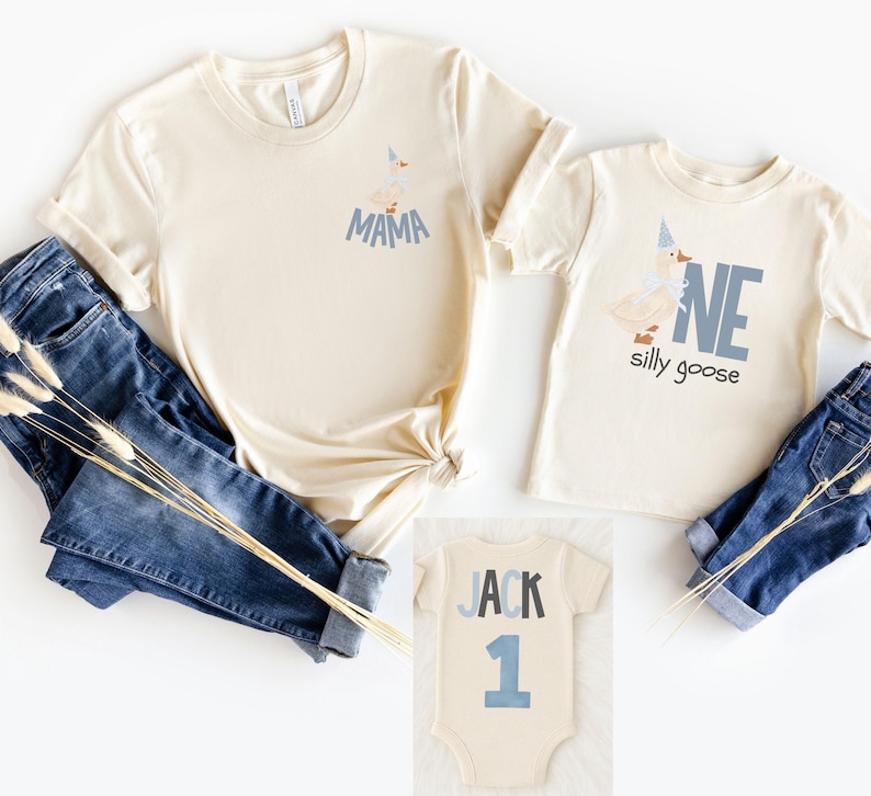 May include: Cream-colored t-shirts and a onesie with blue text and graphics. One shirt says "MAMA" and features a goose graphic. Another shirt says "ONE silly goose". The onesie says "JACK 1". Blue jeans are also in the image.