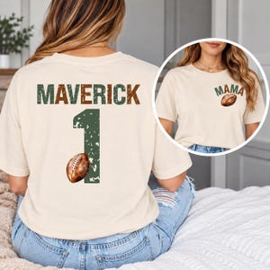 May include: A cream-coloured t-shirt featuring a distressed green and brown graphic on the back that reads MAVERICK above a large number 1 and a football. The front chest area displays the word MAMA in green and brown with a small football.