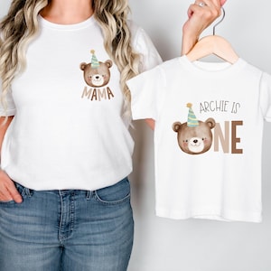 May include: Matching white t-shirts with a brown bear wearing a party hat. The adult shirt says "Mama" and the child's shirt says "Archie is One".