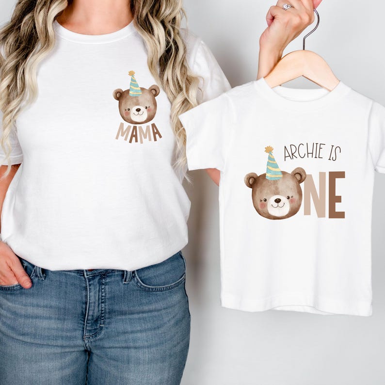 May include: Matching white t-shirts with a brown bear wearing a party hat. The adult shirt says "Mama" and the child's shirt says "Archie is One".