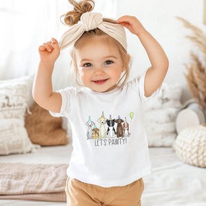 Op de afbeelding: Een jong kind draagt een wit T-shirt met een afbeelding van honden die feestmutsen dragen en de tekst "Let's Pawty!" Het kind draagt een lichtbruin hoofdband en heeft bruin haar.
