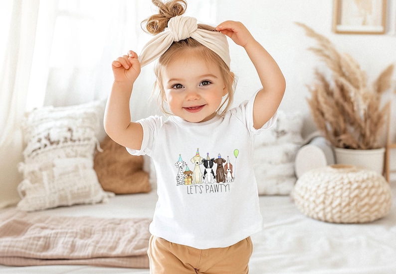Op de afbeelding: Een jong kind draagt een wit T-shirt met een afbeelding van honden die feestmutsen dragen en de tekst "Let's Pawty!" Het kind draagt een lichtbruin hoofdband en heeft bruin haar.