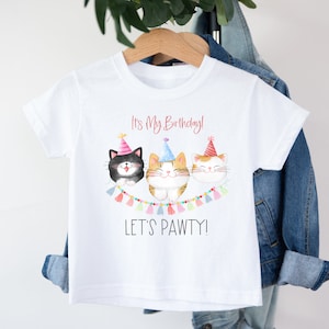 May include: White t-shirt with a birthday theme featuring three cartoon cats wearing party hats and a banner that says "It's My Birthday! Let's Pawty!"