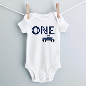 May include: White baby onesie with the word "ONE" in navy blue block letters and a navy blue truck graphic. The onesie is hanging on a clothesline with wooden clothespins.