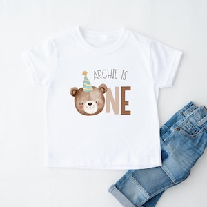 May include: White t-shirt with a brown bear wearing a party hat and the text "Archie is one".