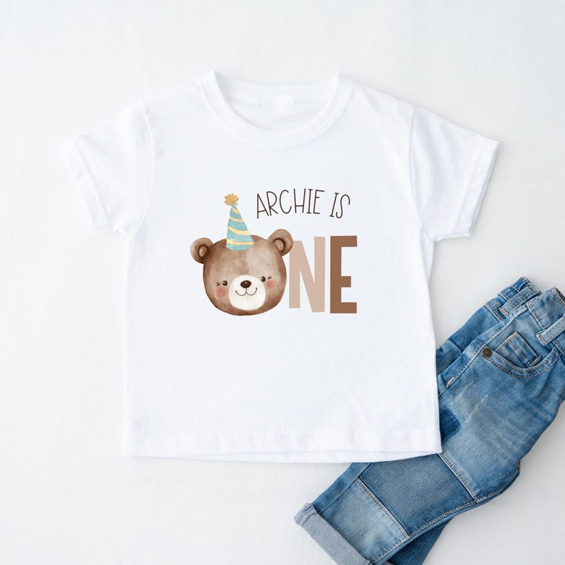 May include: White t-shirt with a brown bear wearing a party hat and the text "Archie is one".