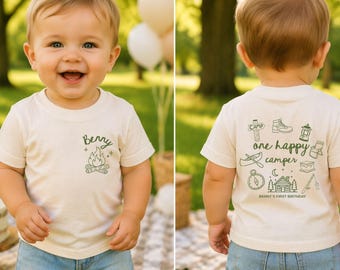 Puede incluir: Una camiseta color crema con temática de camping. La parte delantera de la camiseta tiene el nombre "Berry" encima de un gráfico de fogata. La parte trasera de la camiseta dice "one happy camper" con ilustraciones relacionadas con el camping y "Benny's First Birthday".