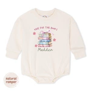 May include: A natural romper with long sleeves. The front features a stack of books with a pink ribbon and the text "ONE FOR THE BOOKS" and "Madden". Pink flower accents surround the book stack. The words "natural romper" are in a circle at the bottom.