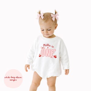 May include: A white long-sleeve romper with the text "Millie is ONE" in red. The word "ONE" is in a pink gingham pattern. The romper has a round neckline and is designed for a baby.