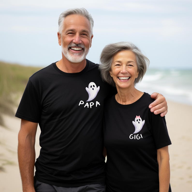 May include: Two people wearing matching black t-shirts with ghost designs. One shirt says "PAPA" and the other says "GIGI". The ghost designs are white with a simple cartoon style. The background is a beach scene.