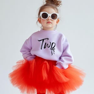 May include: A young child wearing a lavender sweatshirt with the word "Two" in black script, paired with a vibrant red tutu skirt. The child is also wearing white sunglasses.