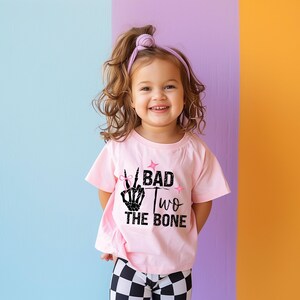 May include: A pink toddler t-shirt with a black skeleton hand making a peace sign and the text "Bad Two The Bone".