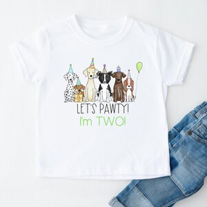 Op de afbeelding: Wit T-shirt met een afbeelding van vijf honden die feestmutsen dragen en een cadeau vasthouden. De tekst "Let's Pawty! I'm Two!" staat onder de afbeelding.