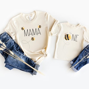 May include: Two cream colored t-shirts with bee graphics. The larger shirt says "MAMA" with three bees. The smaller shirt says "ONE" with a bee.