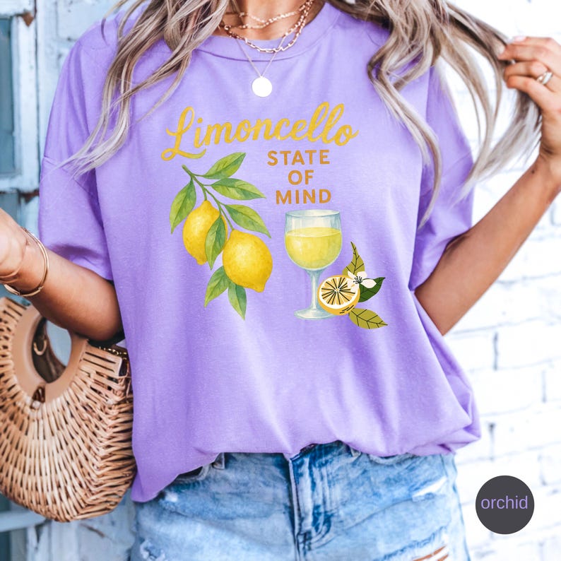 May include: A lilac-colored t-shirt with the words "Limoncello State of Mind" in yellow script. The shirt features a graphic of lemons, leaves, and a glass of limoncello. The word "orchid" is printed in a circle on the bottom right.
