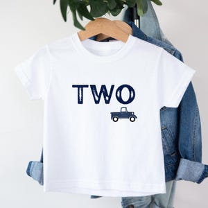 May include: White toddler t-shirt with the word "TWO" in navy blue block letters and a navy blue truck graphic. The shirt is hanging on a wooden hanger.
