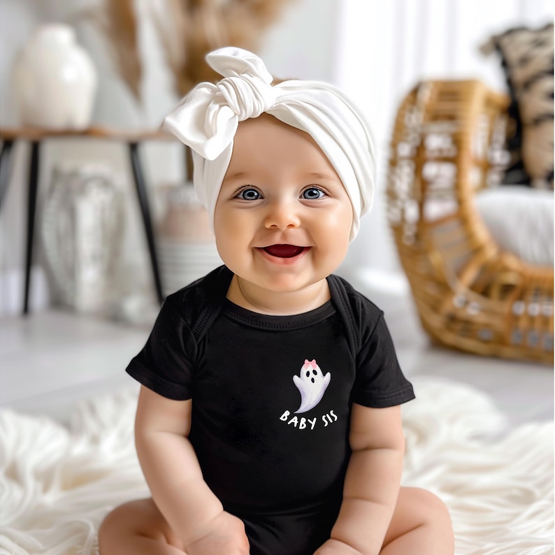 May include: A smiling baby wearing a black onesie with a white ghost graphic and the text "Baby Sis." The baby also wears a white knotted headband. The baby is sitting on a white fluffy rug.