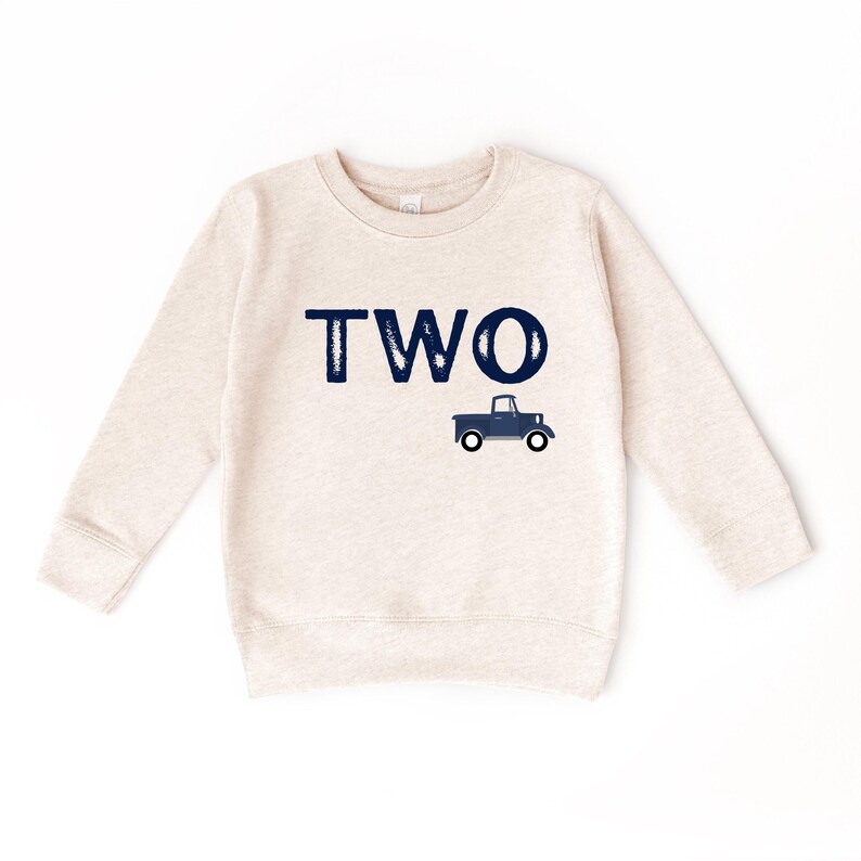 May include: A light beige sweatshirt with long sleeves. The word "TWO" is printed in navy blue, with a navy blue truck graphic below it. The sweatshirt is designed for children.