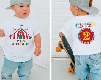 May include: A toddler wearing a white t-shirt with a circus-themed graphic and the words "MAIN ATTRACTION". The back of the shirt has the name "OLIVER" and the number "2". The child is wearing a gray baseball cap and blue jeans.