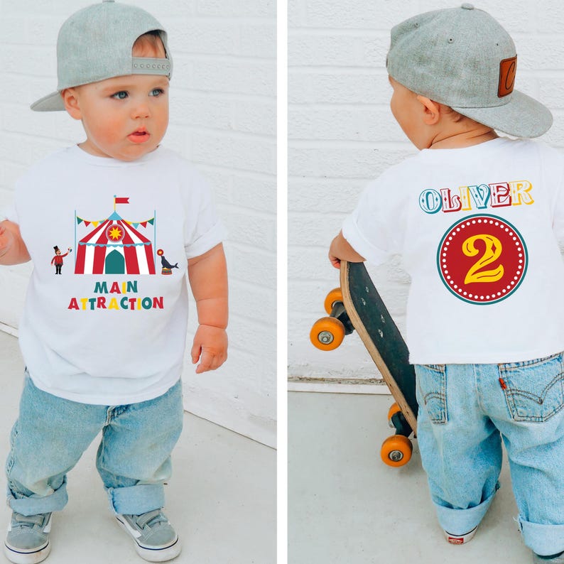 May include: A toddler wearing a white t-shirt with a circus-themed graphic and the words "MAIN ATTRACTION". The back of the shirt has the name "OLIVER" and the number "2". The child is wearing a gray baseball cap and blue jeans.