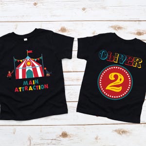 May include: Two black t-shirts with colorful circus-themed designs. One shirt features a circus tent with the words "MAIN ATTRACTION". The other shirt has the name "OLIVER" and the number "2" inside a circle.
