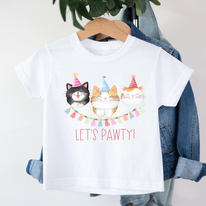 May include: White t-shirt with a graphic of three cats wearing party hats and a banner that says "Let's Pawty!"