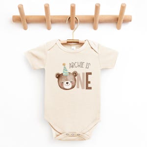 May include: A beige baby onesie with a brown bear wearing a blue and yellow party hat. The onesie has the text "Archie is one" printed on it.