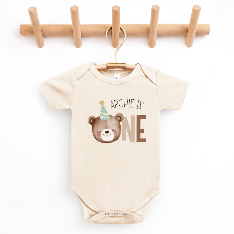 May include: A beige baby onesie with a brown bear wearing a blue and yellow party hat. The onesie has the text "Archie is one" printed on it.