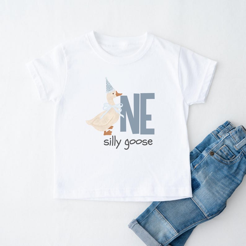 May include: White toddler t-shirt with a whimsical design featuring a goose wearing a party hat and a bow. The text "NE silly goose" is printed in blue. A pair of blue jeans is next to the shirt.