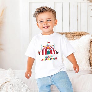May include: A white t-shirt with a colorful circus tent graphic and the words "MAIN ATTRACTION" in rainbow colors. The shirt is worn by a young child wearing blue jeans.
