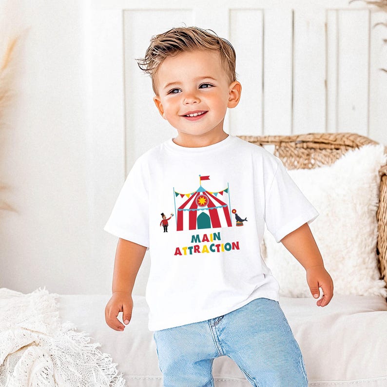 May include: A white t-shirt with a colorful circus tent graphic and the words "MAIN ATTRACTION" in rainbow colors. The shirt is worn by a young child wearing blue jeans.