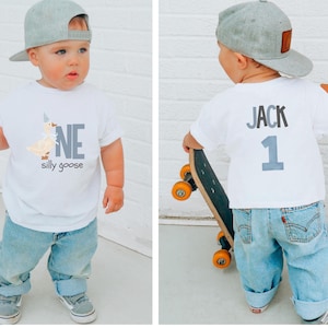 May include: A toddler wearing a white t-shirt with a cartoon goose and the words "ONE silly goose". The child is also wearing light blue jeans, gray sneakers, and a gray baseball cap. The back of the shirt says "JACK 1".
