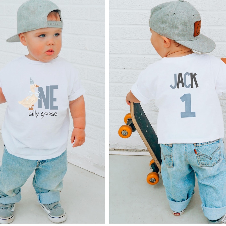 May include: A toddler wearing a white t-shirt with a cartoon goose and the words "ONE silly goose". The child is also wearing light blue jeans, gray sneakers, and a gray baseball cap. The back of the shirt says "JACK 1".