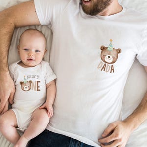 May include: A white onesie with a brown bear wearing a party hat and the text "Archie is one". A white t-shirt with a brown bear wearing a party hat and the text "Dada".