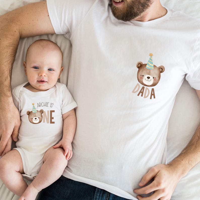 May include: A white onesie with a brown bear wearing a party hat and the text "Archie is one". A white t-shirt with a brown bear wearing a party hat and the text "Dada".