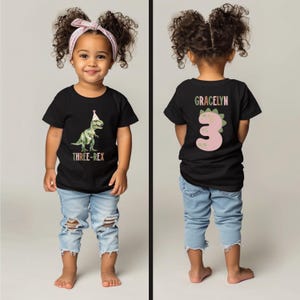May include: Black t-shirt featuring a green dinosaur in a party hat and the text "THREE-REX". The back of the shirt displays the name "GRACELYN" and a pink number "3" with a dinosaur design. The child is wearing ripped blue jeans.