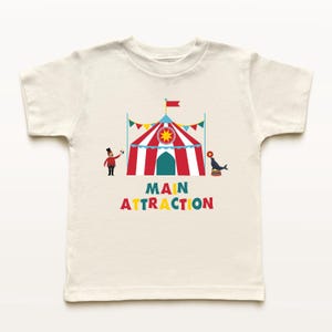 May include: Off-white t-shirt featuring a colorful circus design. The design includes a red and white striped tent, a ringmaster, a seal balancing on a ball, and the words "MAIN ATTRACTION" in colorful letters. A fun, playful design.