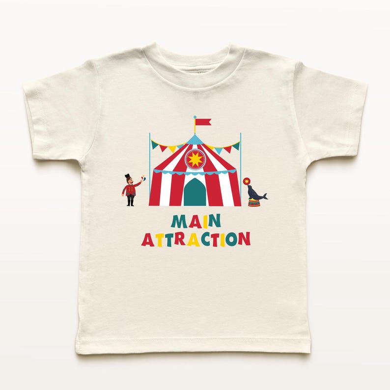 May include: Off-white t-shirt featuring a colorful circus design. The design includes a red and white striped tent, a ringmaster, a seal balancing on a ball, and the words "MAIN ATTRACTION" in colorful letters. A fun, playful design.