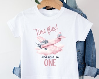 May include: White t-shirt with a pink airplane and clouds graphic. The text "Time flies! and now I'm ONE" is printed on the shirt.
