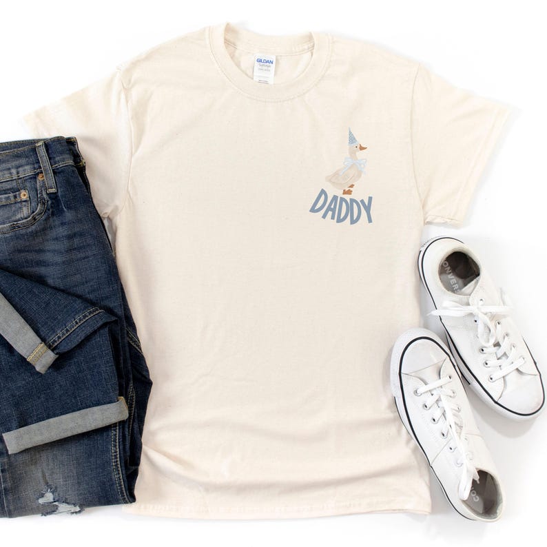May include: Cream-colored t-shirt with a cartoon goose wearing a party hat and bow tie, with the word "DADDY" in blue. The shirt is paired with blue jeans and white sneakers.