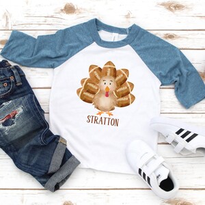 May include: A white and blue baseball style shirt with a cartoon turkey wearing footballs for feathers. The shirt has the name "Stratton" printed on the front.