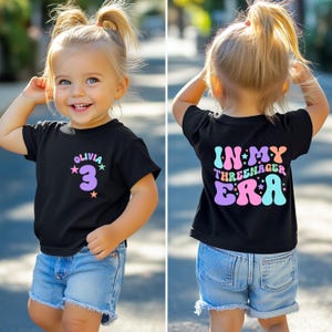 May include: A black t-shirt with a colorful "In My Three-nager Era" graphic on the back and a purple number "3" with stars on the front. The shirt is being worn by a child.